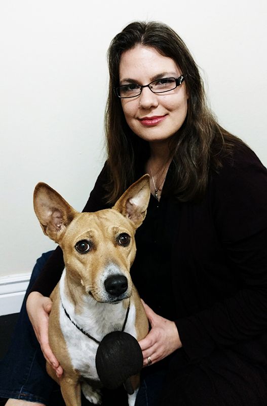 A woman with glasses smiles while sitting next to a dog with pointy ears, both facing the camera.