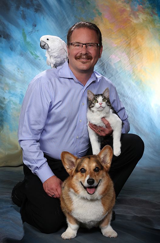 Man smiling with a parrot on his shoulder, holding a cat, and a corgi sitting by his side.